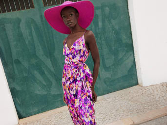 Woman in a sleeveless, patterned wrap dress stands in front of a green house wall | C&A