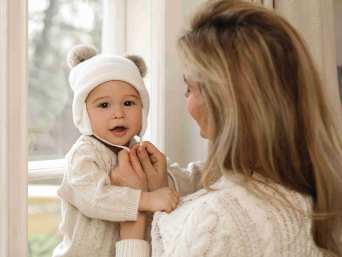 Frau bindet einem Baby eine große Babymütze mit Ohrenklappen unter dem Kinn zu Frau bindet einem Baby eine große Babymütze mit Ohrenklappen unter dem Kinn zu