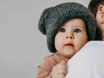 Mann hält Baby mit großer, grauer Beanie, deren Mützengröße dem Baby offensichtlich nicht passt Mann hält Baby mit großer, grauer Beanie, deren Mützengröße dem Baby offensichtlich nicht passt