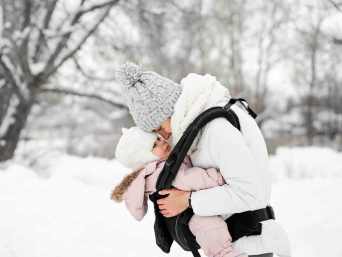 Frau in weißer Winterkleidung trägt ein dick eingepacktes Baby in der Babytrage im Schnee – ein Beispiel, wie man ein Winterbaby anziehen kann