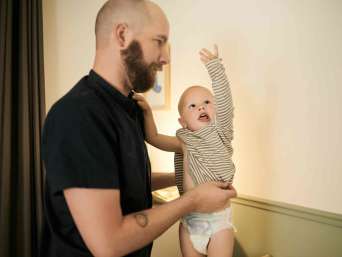 Mann mit Bart zieht Baby ein langärmeliges Unterhemd an – Symbolbild für das Thema "Ab wann keine Bodys mehr?" Mann mit Bart zieht Baby ein langärmeliges Unterhemd an – Symbolbild für das Thema "Ab wann keine Bodys mehr?"
