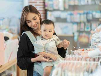 Frau mit Baby in Trage überlegt in einem Laden, wann ein Unterhemd statt Body besser ist. Frau mit Baby in Trage überlegt in einem Laden, wann ein Unterhemd statt Body besser ist.