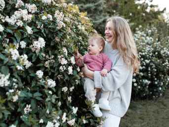 Blonde Frau zeigt Baby in leichter Babykleidung im Frühling weiße Blüten