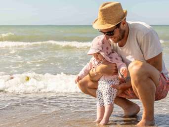 Mann mit Sonnenhut hält am Strand vorsichtig die Füßchen eines Babys in Sommerkleidung ins Meer. Mann mit Sonnenhut hält am Strand vorsichtig die Füßchen eines Babys in Sommerkleidung ins Meer.