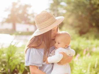Frau mit Sonnenhut küsst Baby in langärmeliger Babykleidung im Sommer in einem Garten. Frau mit Sonnenhut küsst Baby in langärmeliger Babykleidung im Sommer in einem Garten.