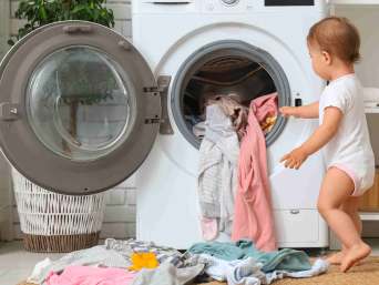 Baby steht vor dem Babykleidung Waschen, vor einer geöffneten Waschmaschine voller Kleidung Baby steht vor dem Babykleidung Waschen, vor einer geöffneten Waschmaschine voller Kleidung