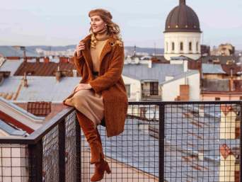 Blonde Frau mit Mantel, Kleid und Stiefeln vor Stadtkulisse – zeigt, wie man ein Kleid im Winter kombinieren kann.