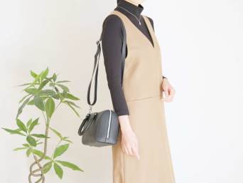 Frau mit schwarzem Rollkragenpullover unter beigem Trägerkleid steht neben einer Topfpflanze.