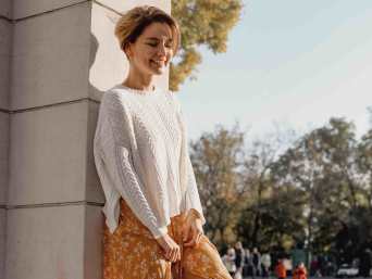 Lachende blonde Frau mit weißem Pulli über dem Kleid lehnt an einer Säule im Freien.