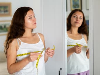 Calculer taille soutien-gorge : une jeune femme se tient devant un miroir avec un mètre ruban.