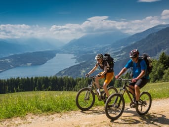 Cómo preparar un viaje en bici: dos ciclistas en ruta con las vistas de un lago de fondo.