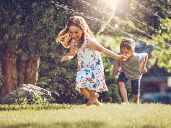 Zwei Kinder laufen lachend durch den Wasserstrahl eines Rasensprengers.