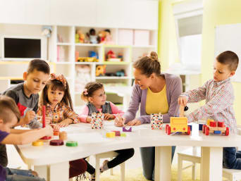 Nástup do školky: skupinka dětí s učitelkou si hraje u stolu.