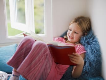 Geschenk 7-Jährige: Kleines Mädchen sitzt an ihrem Fenster und liest in einem Buch.