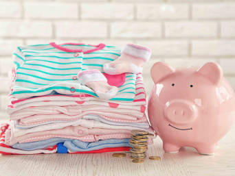 Kosten für die Baby-Erstausstattung – Ein Stapel Kleidung mit einem Sparschwein. 