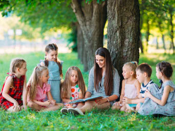 Lesen lernen: Erzieherin liest mit Kindern im Park. 