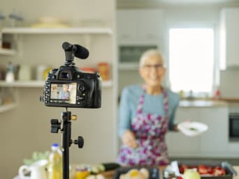 Ouderen online – Oudere vrouw staat voor de camera en draait een kookvideo. 