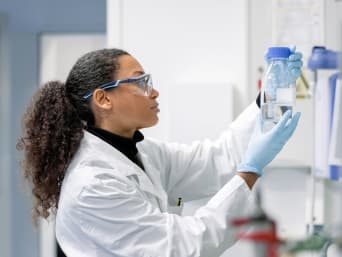 Textillaborant bei der Arbeit: Junge Frau mit Schutzbrille betrachtet im Labor eine Flüssigkeit.