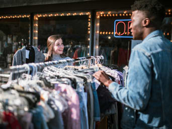 Tweedehands kleding tips: Twee vrienden neuzen buiten voor de winkel tussen vintage kleding aan een kledingrek met tweedehandskleding.