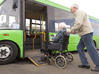 Viaggi per disabili anziani: uomo porta la moglie in carrozzina su un bus.