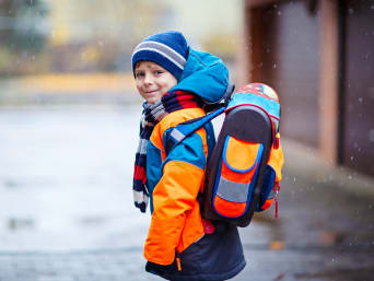 Kleiner Junge in leuchtender Jacke und mit Reflektoren ausgestattetem Schulranzen auf dem Schulweg.