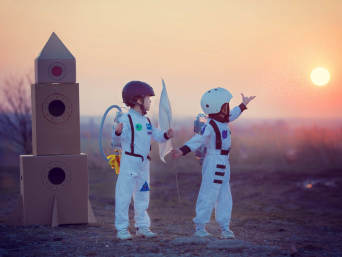 Dwoje dzieci przebranych za astronautów bawi się kartonową rakietą.