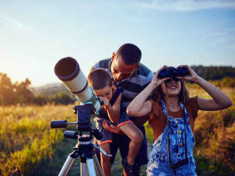Astronomia dla dzieci: tata z synem i córką obserwują niebo za pomocą teleskopu i lornetki.
