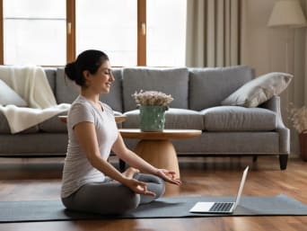 Meditatie voor beginners: jonge vrouw zit thuis op een yogamat en neemt deel aan een online meditatieles.
