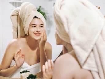 Domowe maseczka na twarz: kobieta z ręcznikiem na głowie nakłada na twarz własnoręcznie zrobioną maseczkę.