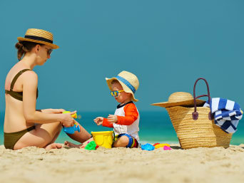 Protección solar para niños: un niño pequeño con la ropa adecuada para protegerse del sol juega en la playa con su madre.