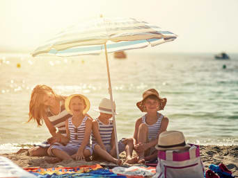Jak chronić skórę przed słońcem?  – kobieta z dziećmi na plaży pod parasolem przeciwsłonecznym.