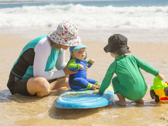 Packliste Sommerferien – Mutter, Baby und Kleinkind mit Sonnenhüten spielen am Strand.