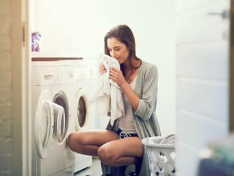 Jak pozbyć się zapachu potu z ubrań? Kobieta kuca przed pralką i wącha pranie. 