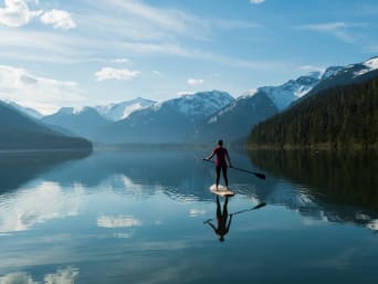 Stand up paddling – Een vrouw supt over een rustig meer, omgeven door besneeuwde bergen.