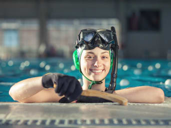 Unterwasserhockey: Spielerin stützt sich am Beckenrand ab und hält ihren Schläger in die Kamera.