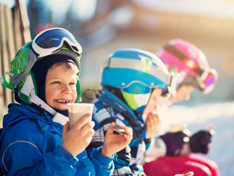 Vacanze sulla neve con bambini: bambino con tuta da sci fa una pausa su una pista da sci per bambini.