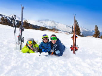 Skifahren Regeln: Familie liegt im Schnee und lacht in die Kamera.