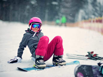 Snowboard fahren lernen: Kind sitzt auf dem Boden und übt mit dem Snowboard aufzustehen.