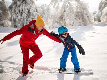 Jak nauczyć dziecko jazdy na snowboardzie – maluch z trenerem podczas pierwszej lekcji na stoku. 