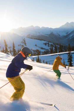 Schönste Skigebiete: Skifahrer fahren im Sonnenschein einen schneebedeckten Hang herunter.