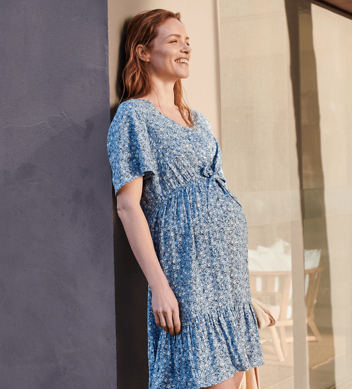 Těhotná žena si užívá slunce v květovaných těhotenských šatech s nabíráním nad břichem.