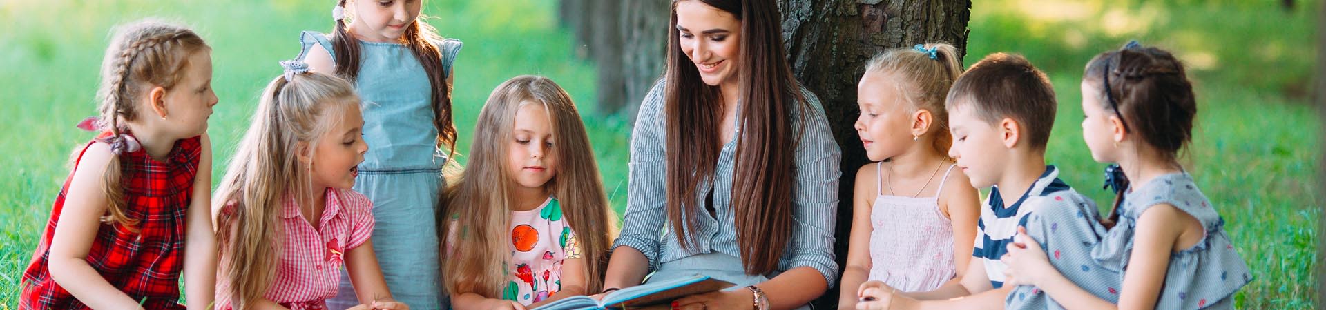 Lesen lernen: Erzieherin liest mit Kindern im Park.