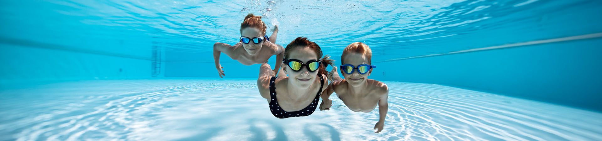 Three children with swimming goggles dive in the pool.