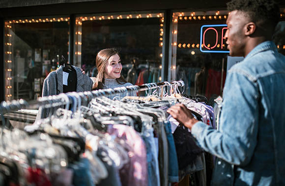 Twee vrienden neuzen buiten voor de winkel tussen vintage kleding aan een kledingrek met tweedehandskleding.