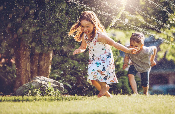 Zwei Kinder laufen lachend durch den Wasserstrahl eines Rasensprengers. Zwei Kinder laufen lachend durch den Wasserstrahl eines Rasensprengers.