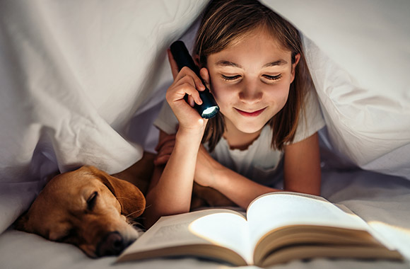 Une fille lit un livre sous sa couverture à l’aide d’une lampe torche.