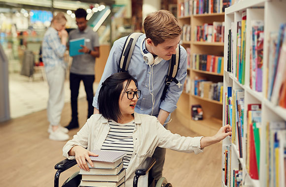 Studentka na wózku razem z asystentem szuka materiałów w bibliotece uniwersyteckiej. Studentka na wózku razem z asystentem szuka materiałów w bibliotece uniwersyteckiej.