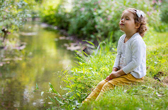 Klein meisje zit bij een rivier met haar ogen dicht en luistert naar het klaterende water.