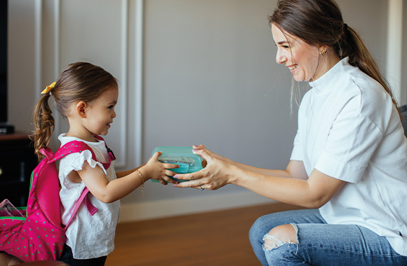 Kindergarten Checkliste: Mutter gibt ihrer Tochter die Brotdose für den Kindergarten. Kindergarten Checkliste: Mutter gibt ihrer Tochter die Brotdose für den Kindergarten.